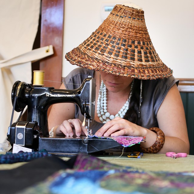 A woman wearing a traditional woven hat sews colorful material