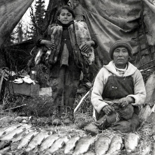 Woman and child in front of tent with fish and furs in foreground.