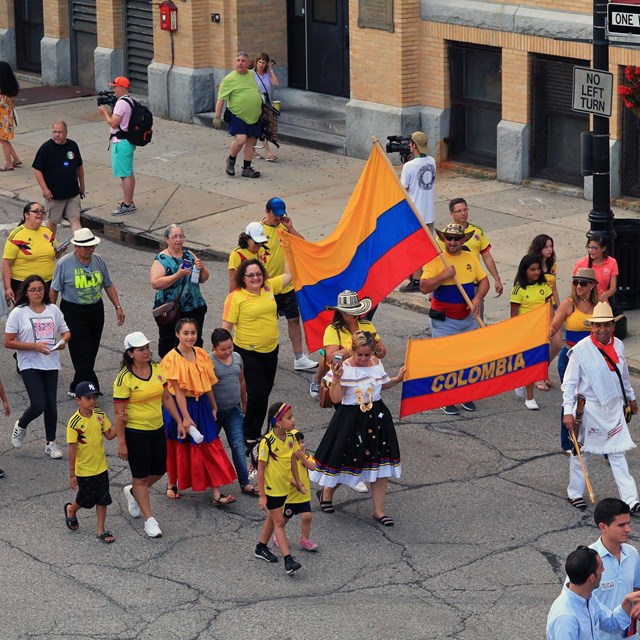 Pedestrians march down the street and on the sidewalk for the parade. A group holds two Colombian fl