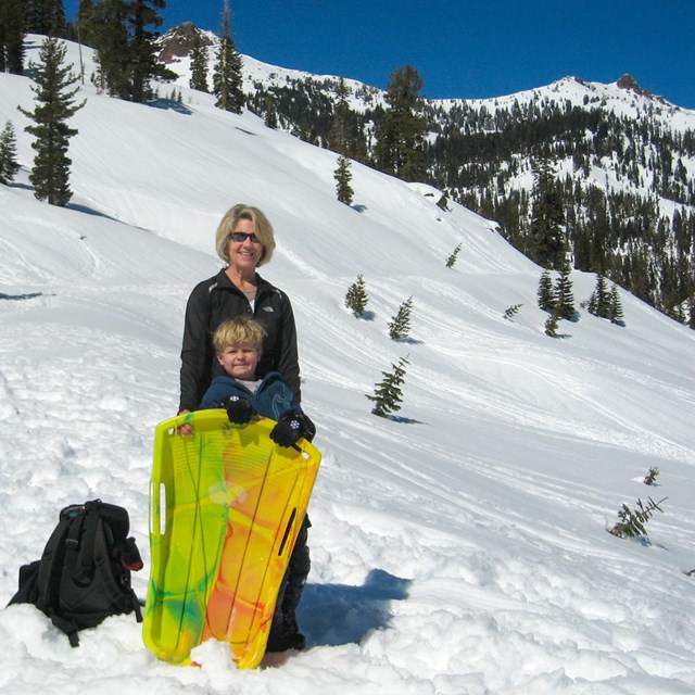 A woman and young boy stand with a yellow sled in a snowy mountain landscape.