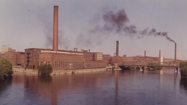 A line of Lowell's mills pouring black smoke into the air with canals in the foreground