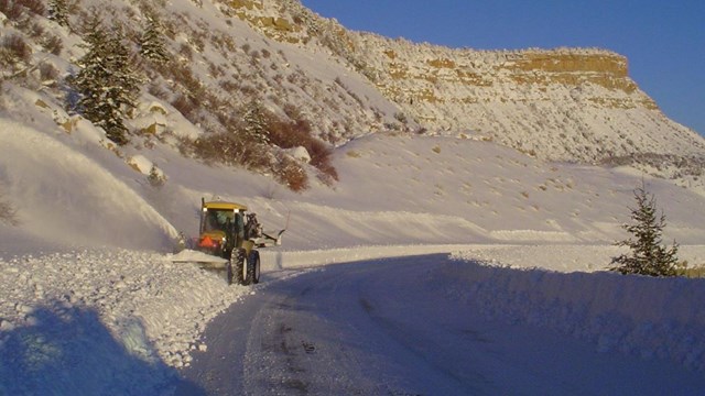 A yellow plow pushes several feet of snow off a road partly in shadow by a mesa cliff wall.
