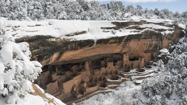 An ancestral stone-masonry village tucked into a natural cliff alcove surrounded by snowy forest