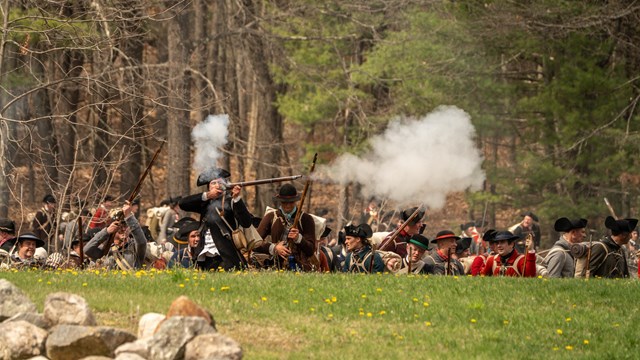 Militia soldiers crouch behind a stone wall loading and firing muskets while surrounded by smoke.
