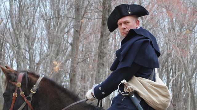 Man in colonial clothing, black cocked hat, dark blue coat and a sword sits on a brown horse.