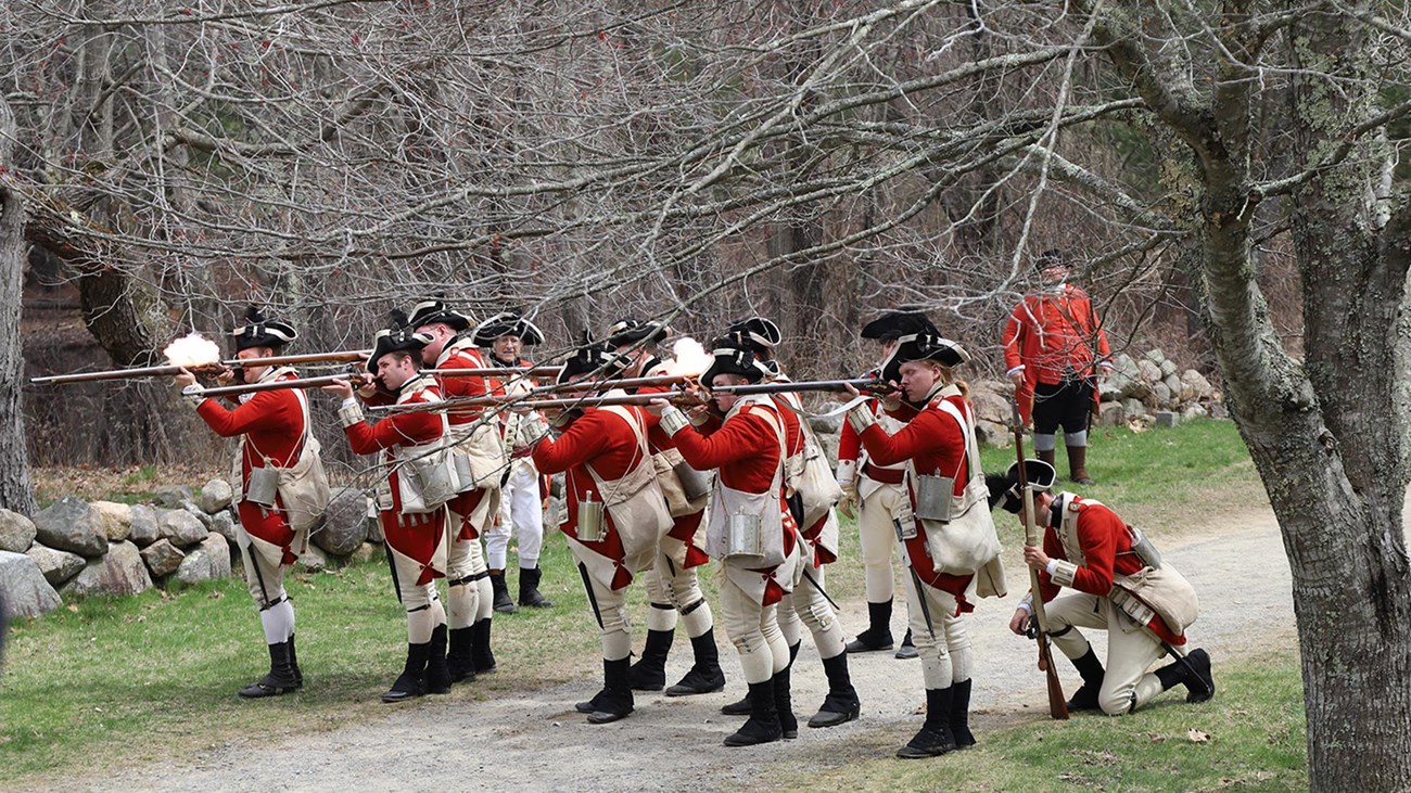 A group of British Soldiers stand on a dirt road aiming their muskets and firing.