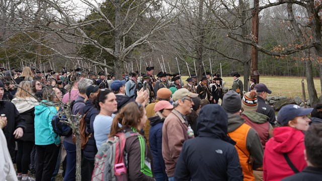A large crowd watches reenactors march along a wooded road.