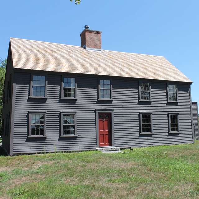 A two story wooden colonial house that is five bays wide sits in a field surrounded by green grass.