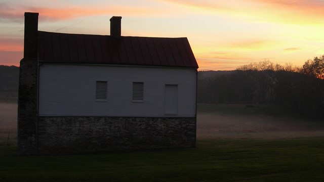 A white structure supported by bricks sits in an open field with the sun setting in the distance
