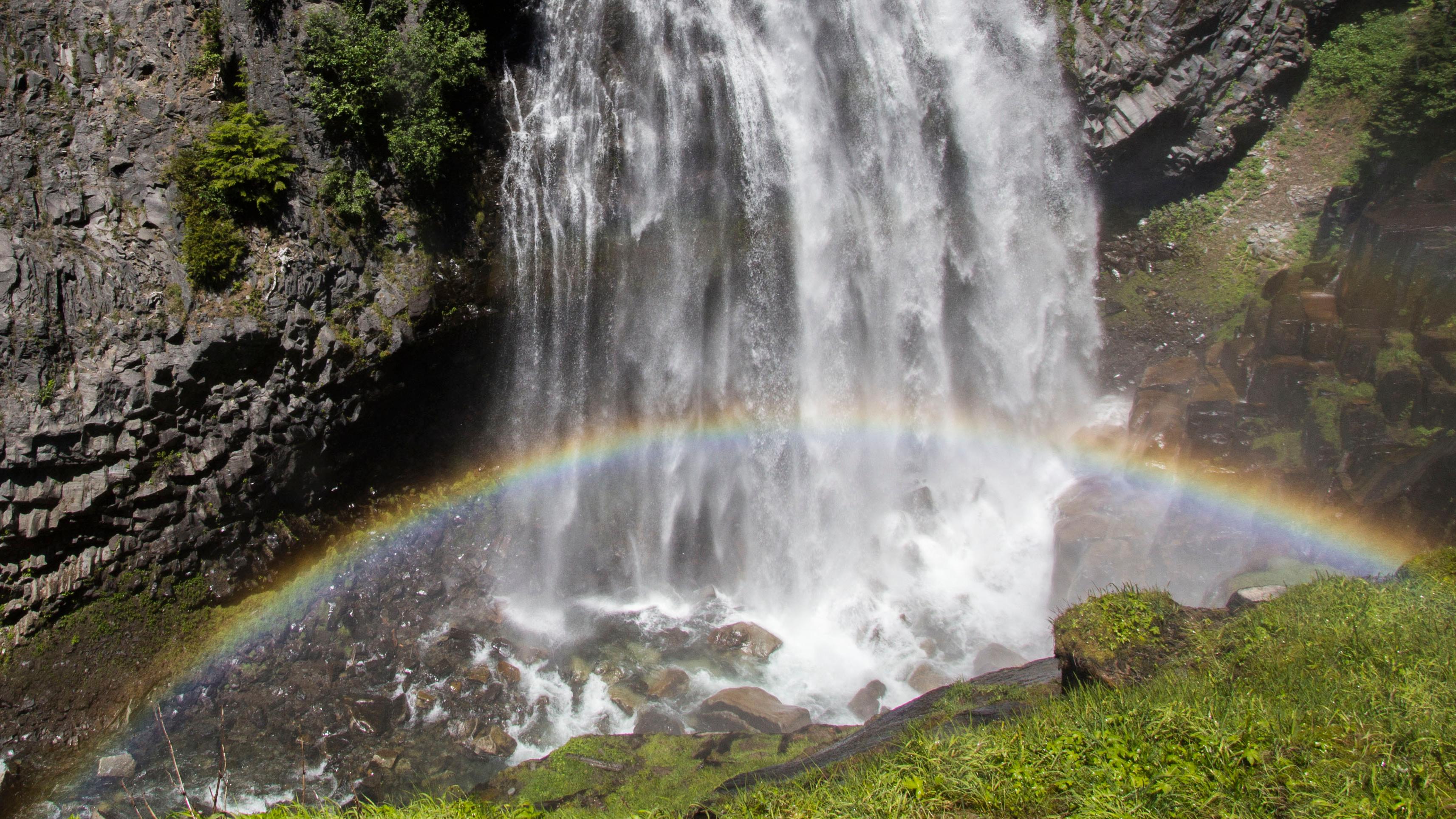 A rainbow arches through the spray of a waterfall cascading down a rocky cliff. 
