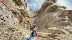 A Mount Rushmore ropes team member working near the chins of Abraham Lincoln and Theodore Roosevelt.