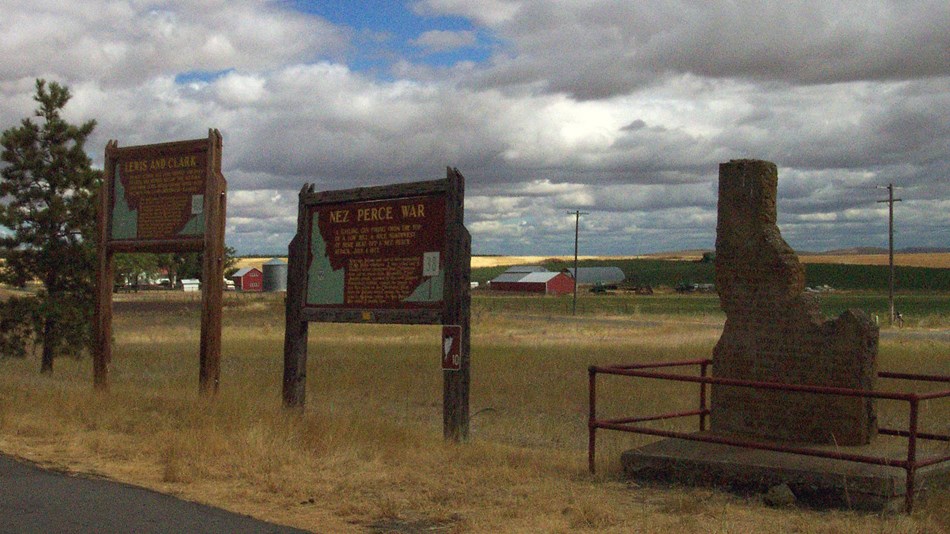Two brown signs and a monument in the shape of Idaho on the side of the road.