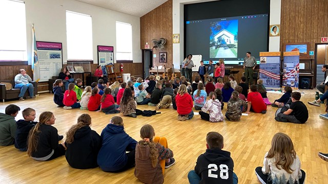 A large group of young students sit on the wooden floor of a large hall facing people speaking.