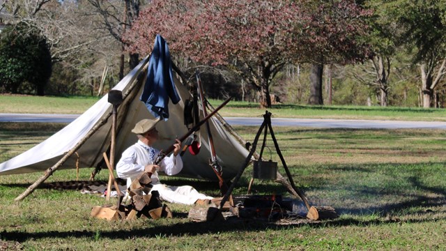 A colonial living historian cleans his musket in front of his tent and the park sign.