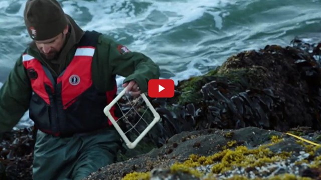 A man in a green uniform and red life jacket holding a piece of field equipment made out of PVC pipe