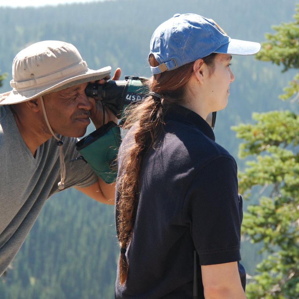 A man looks through a scope. A woman stands next to him.