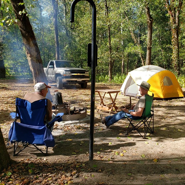 Two gentlemen next to a small fire ring at a campsite, tents and a truck are in the background