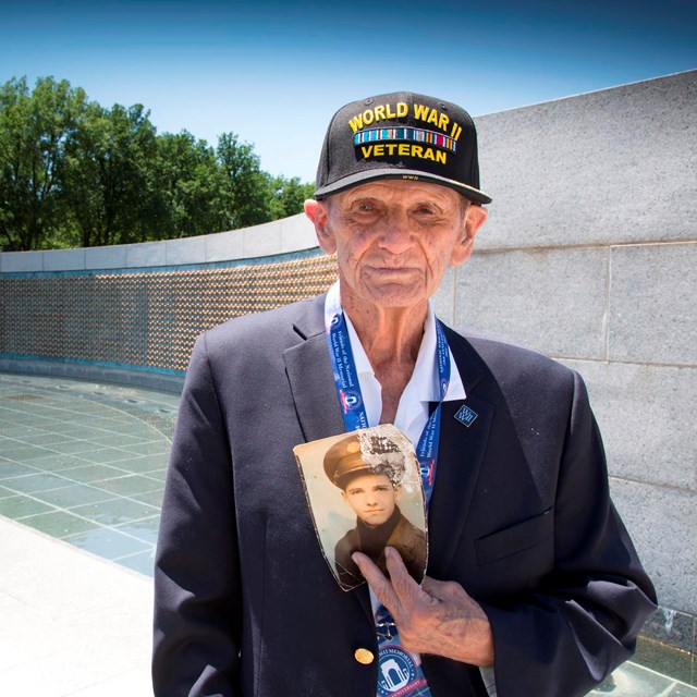 World War II Veteran holding an older photo of himself while standing at the WWII Memorial