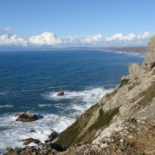 An ocean stretches to the left of a rocky cliff, beyond which is a long beach and clouds.