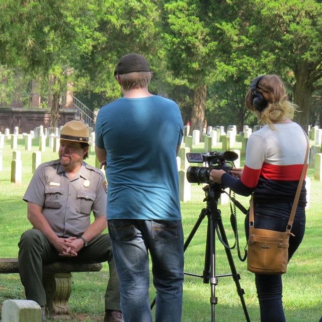 Two people point a video camera at a park ranger on a bench in a cemetery.