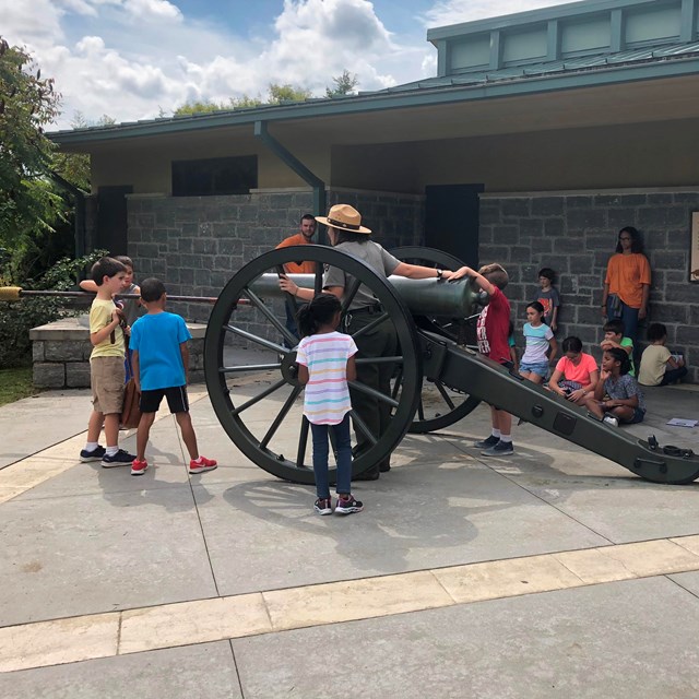 A park ranger stands next to a cannon as a group of students mime loading a cannon.