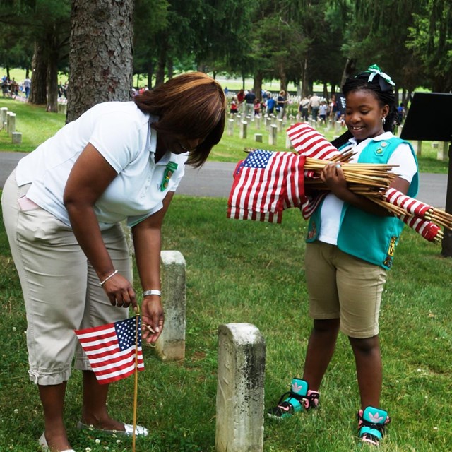A girl scout holds small American Flags. A woman beside her places one flag in front of a headstone.
