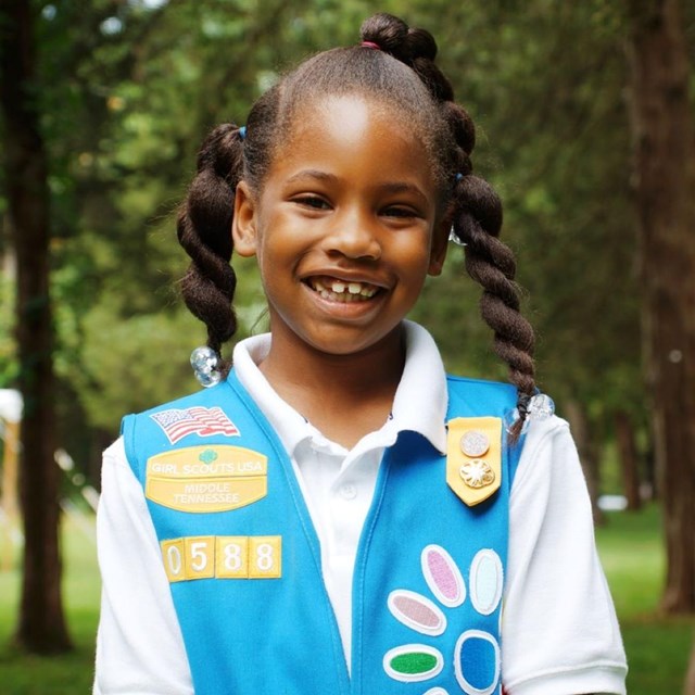 A girl scout stands in front of some trees.