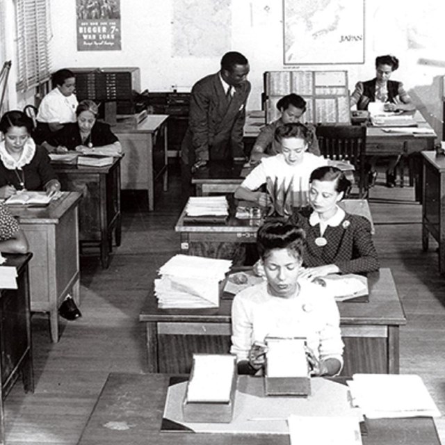 Women sitting in front of desks. An African American male is helping a student sitting in the back.