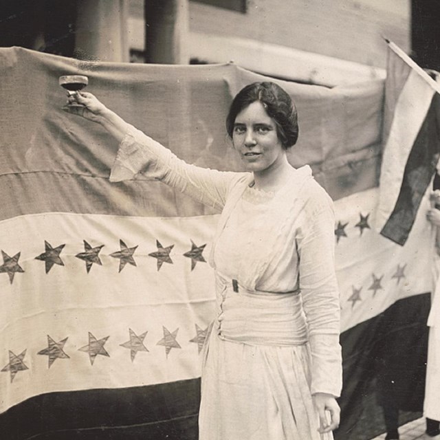 Photo of woman holding a cup in front of a flag. 