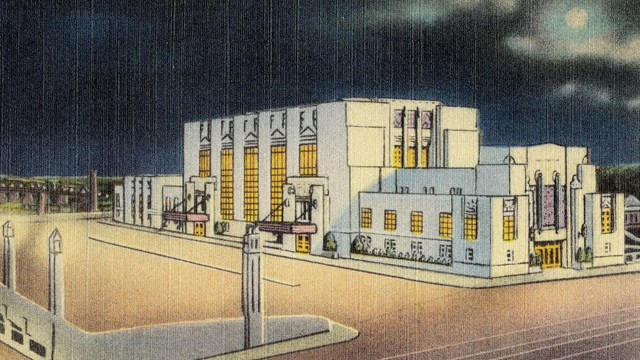 Art-deco train station. Large vertical windows lit up. Fronted by parking area.