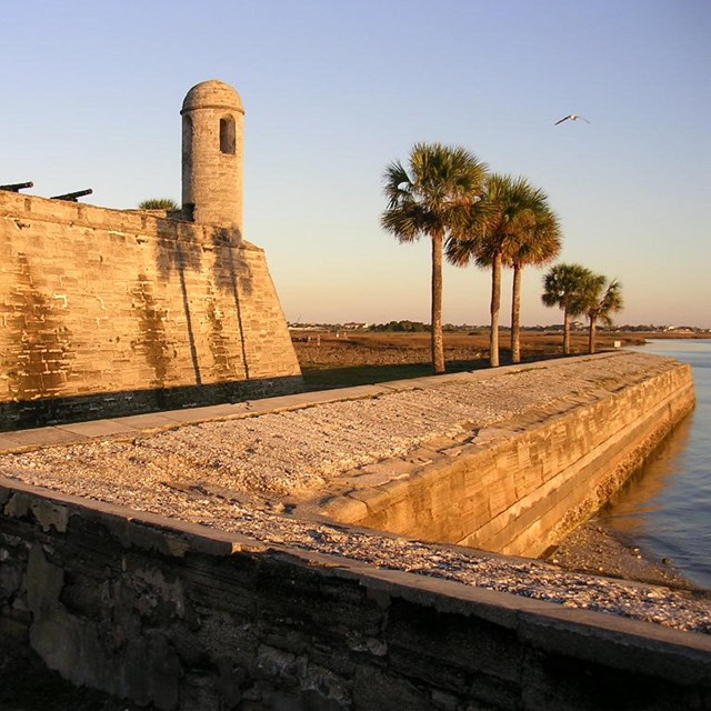 Sunset casts over a stone wall behind a moat. Munitions are mounted behind the wall.