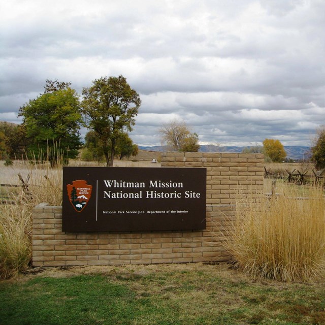Entrance sign reading Whitman Mission National Historic Site, tall grasses beside it