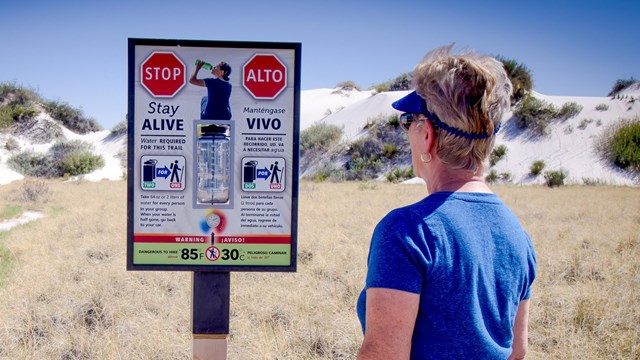 A visitor faces away reading safety sign about bringing water when hiking in the park.