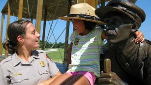 color photo of ranger on left and young girl on right wearing a ranger hat next to a statue of a man