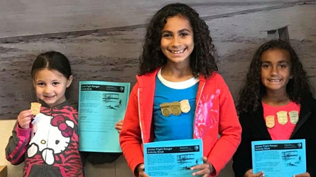 color photo of three girls standing holding blue activity books wearing wooden badges