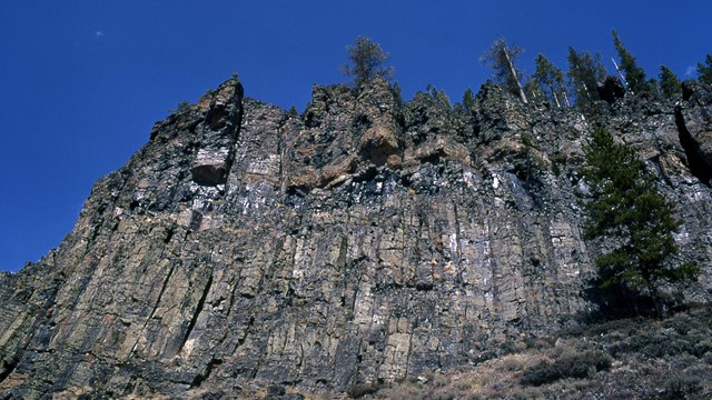 Brown and gray columns of rock make up a cliff that towers up to a deep blue sky.