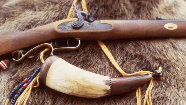 Rifle and powder horn with a map etched on side resting on fur.