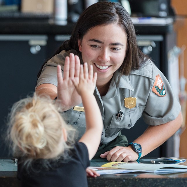 A child with a woman gives a high 5 to a park ranger across a desk.
