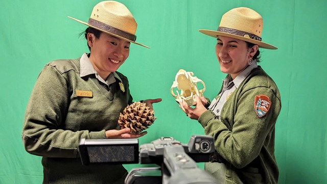 Two park rangers holding props in front of a green screen talking to the camera