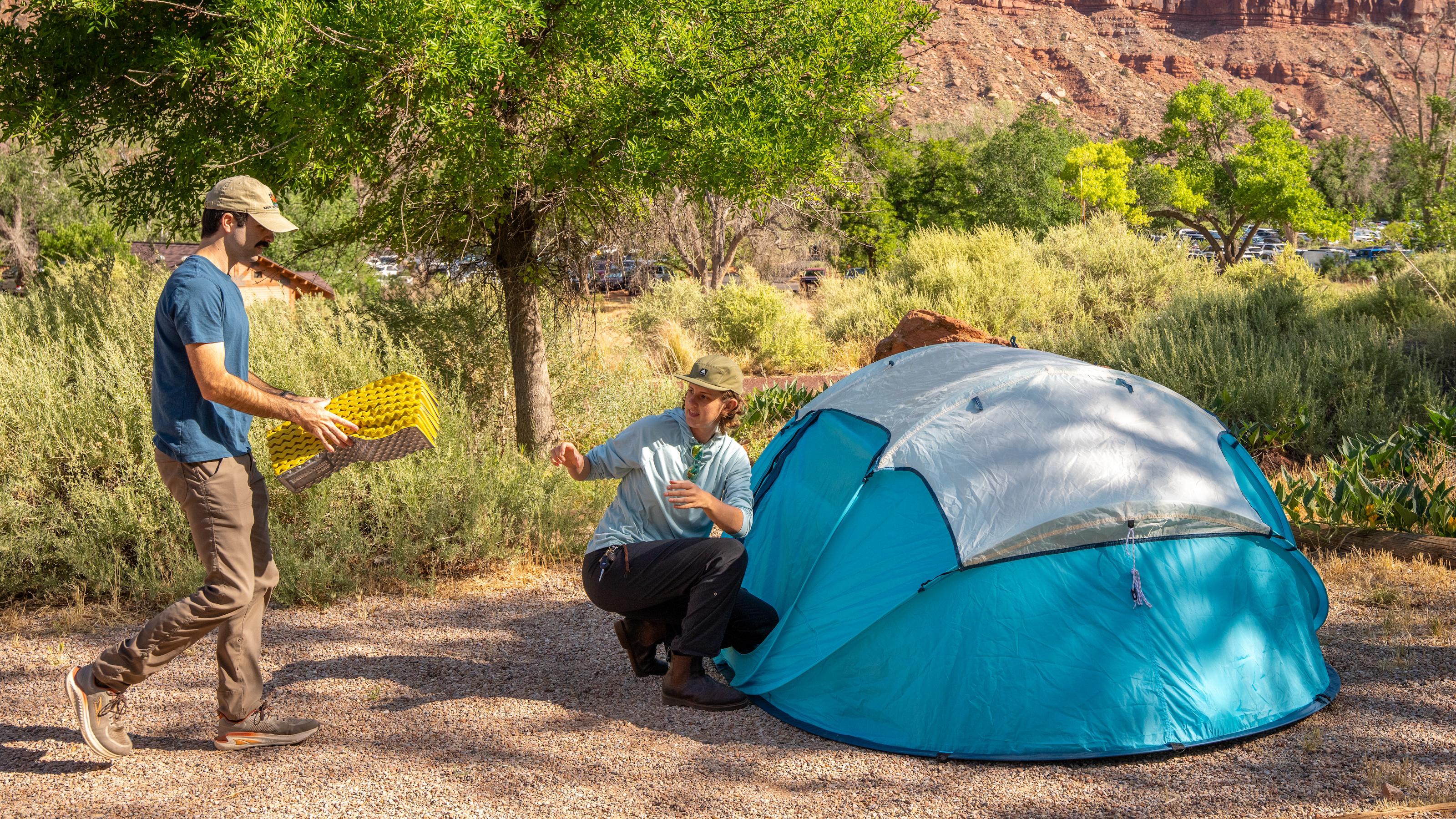 Two people set up camp next to a bright blue tent. Huge sandstone cliffs stand in the background.