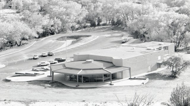 Black and white historic image of building with 1950s cars in front lot and trees in the distance.