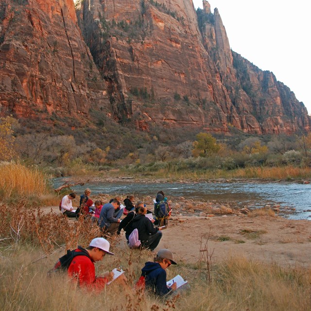Students writing in notebooks along a river bank