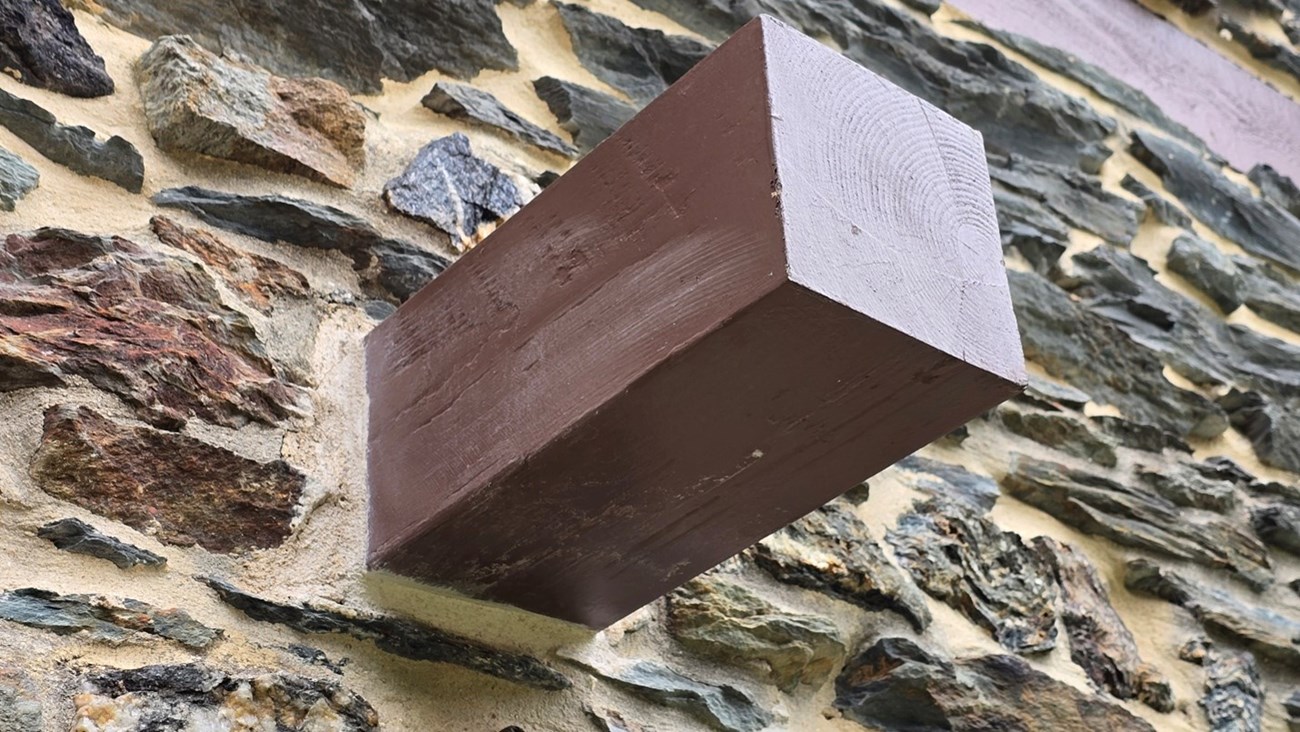 A solid wooden joist, painted brown, sticks out of a stone wall.