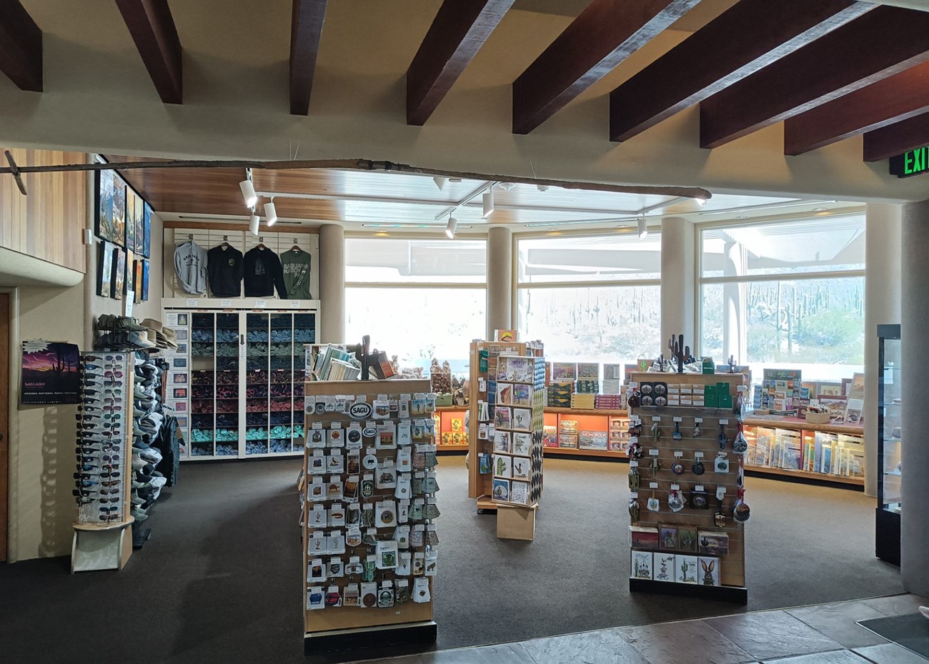 Looking into a clean gift shop. 3 large windows on the back wall show the desert, with items and books for sale at their base. Three main stands create isles in the store, stocked with stickers, patches, postcards, and more. Shirts and hats on the left