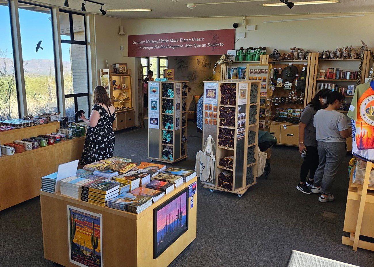 Looking into a clean gift shop. 3 large windows on the back wall show the desert, with items and books for sale at their base. Three main stands create isles in the store, stocked with stickers, patches, postcards, and more. Shirts and hats on the left