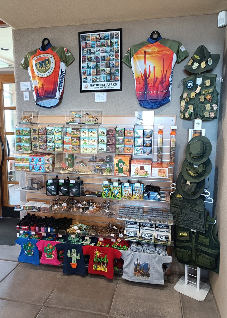 Looking at a tall wall full of merchandise. The top of the wall has two bright colored bike jerseys sandwiching a poster of park images. The shelves below that are full of toys, games, and kids' shirts. To the right is a stand with Jr. Ranger gear.