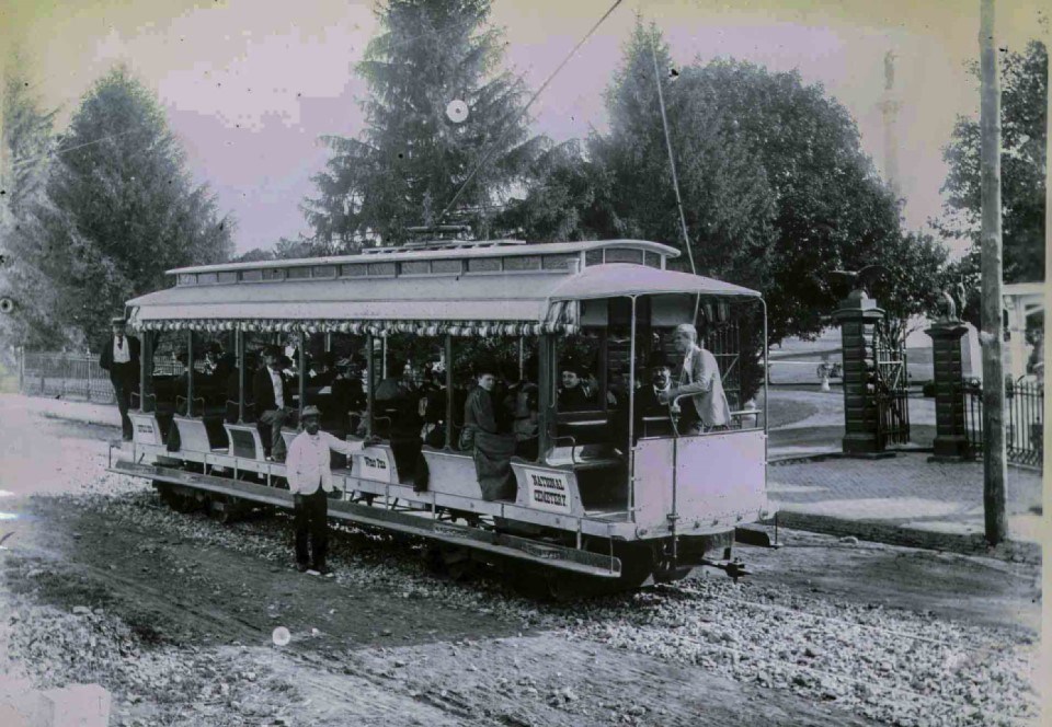 A black and white picture of several men and women positioned inside an electric trolley with one man standing outside of the trolley. Trees, two monuments, and an open gate leading into a cemetery can be seen in the background.