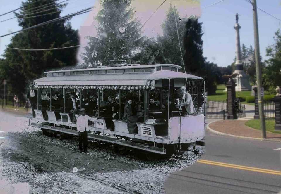 A black and white picture of several men and women positioned inside an electric trolley with one man standing outside of the trolley. Trees, two monuments, and an open gate leading into a cemetery can be seen in the background.