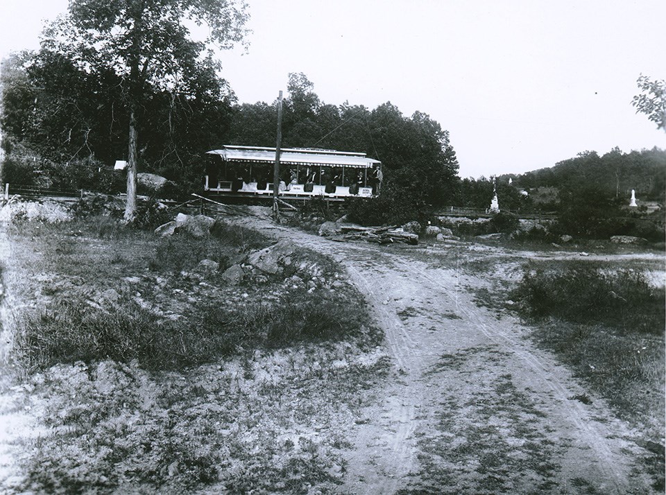 Historical black and white photograph of a trolley running through the battlefields with monuments on both sides in the distance.