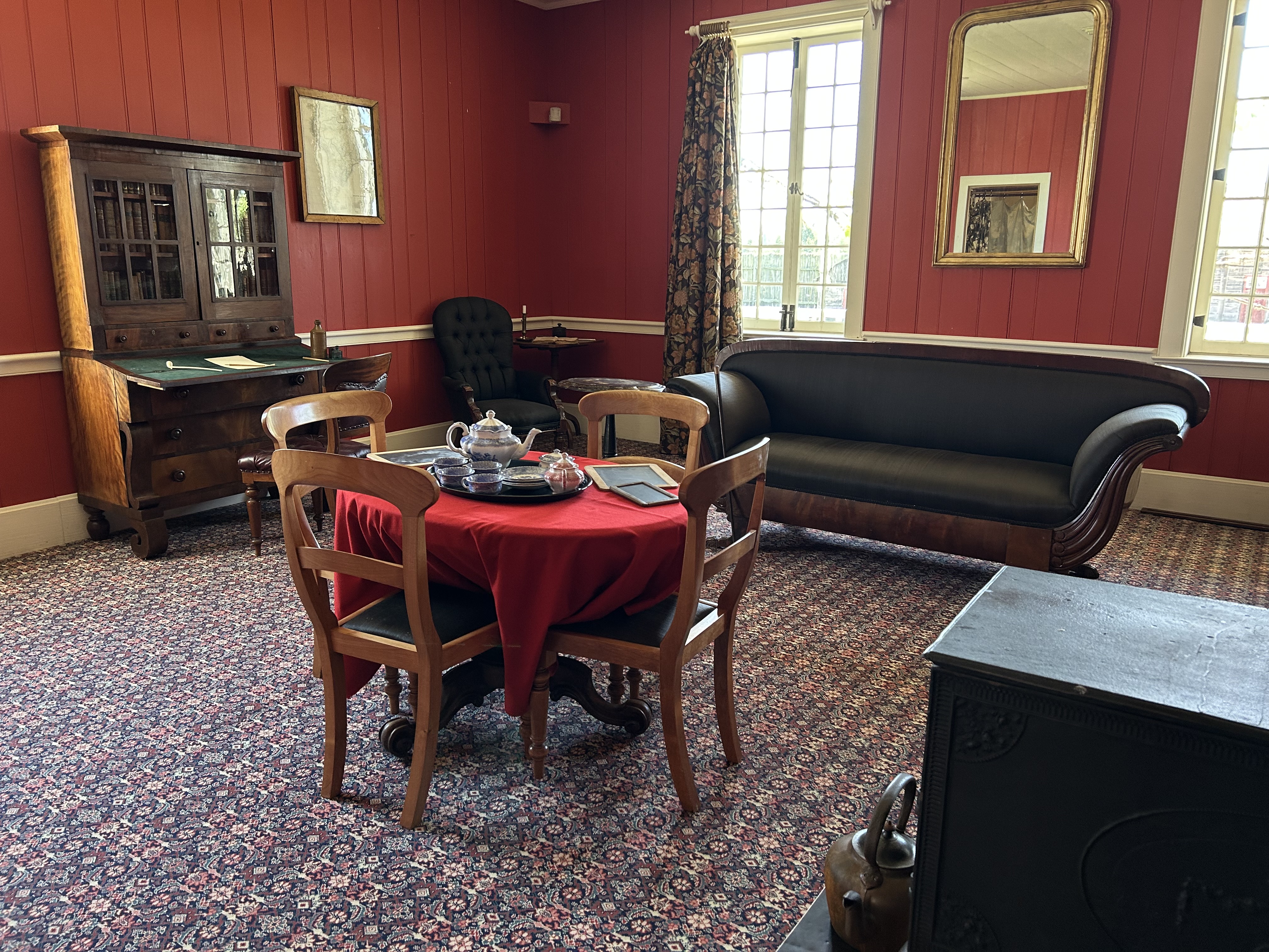 The Douglas Sitting Room in the Chief Factor's House at Fort Vancouver. The room's walls are painted red. Furniture includes a horsehair sofa, table with a tea set, and a bookcase and desk.